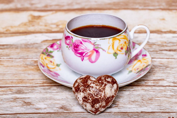 Una taza de porcelana blanca en un platillo con un delicado patrón de rosas rosadas y amarillas con café instantáneo sobre un fondo de madera con un pan de jengibre de chocolate en forma de corazón
. - Foto, imagen