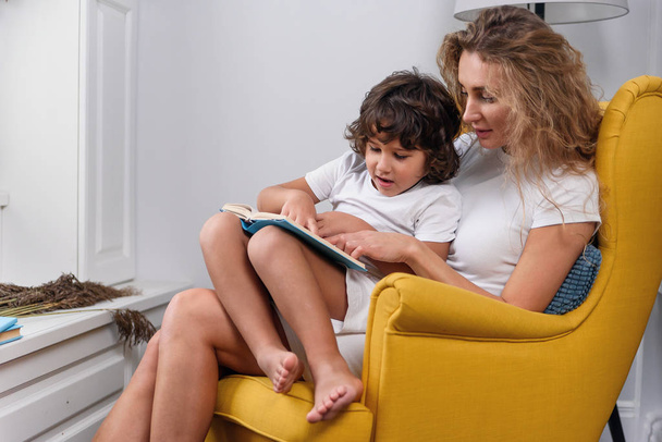 Close up of appealing young concentrated mom which sitting in soft green chair with her 5-aged happy cheerful son and enjoying of reading interesting book - Foto, afbeelding