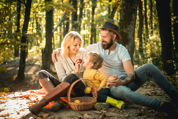 Basket with picnic meal and toys for the kid. Happy family of three lying in the grass in autumn. The concept of a happy family. Young smiling family doing a picnic on an autumns day. - Photo, Image