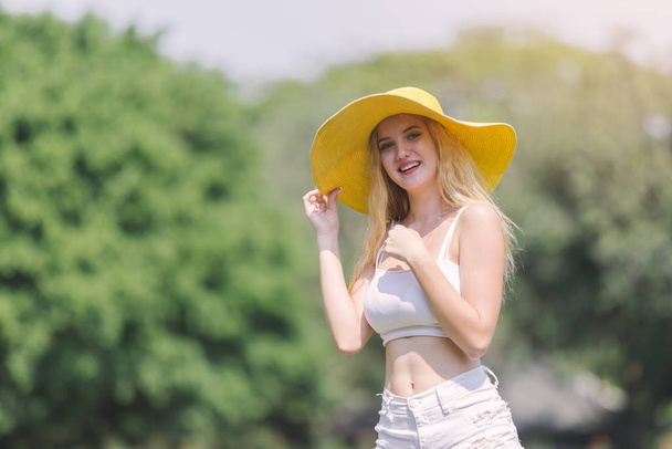 portret mooie jonge vrouw blond haar glimlach met gele hoed poseren buiten in groene tuin. - Foto, afbeelding
