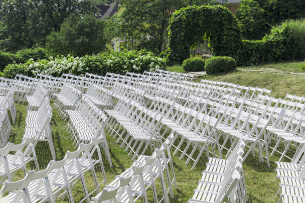 Vue des rangées de chaises blanches en bois, placées en demi-cercle, sans spectateurs, sur fond d'arbustes à fleurs
 - Photo, image