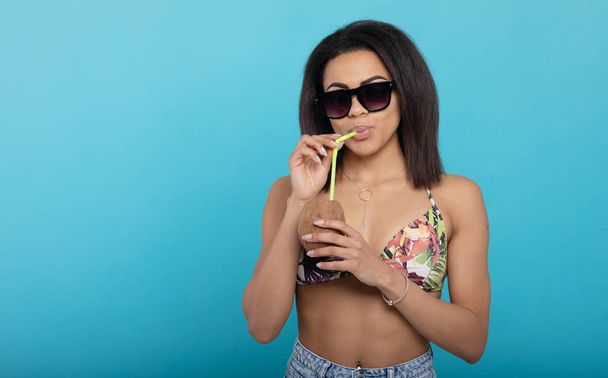 attraktives schwarzes Mädchen in Badebekleidung und Sonnenbrille mit Sommercocktail und lächelt in die Kamera, blauer Hintergrund - Foto, Bild