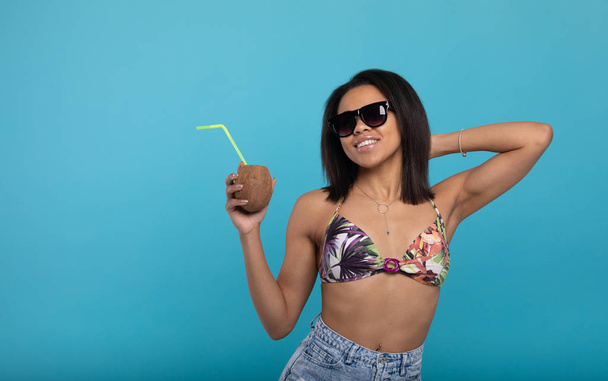 Приваблива чорна дівчина в купальнику і сонцезахисних окулярах тримає літній коктейль і посміхається на камеру, синій фон
 - Фото, зображення