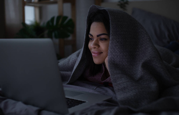 Beautiful young mixed race woman lying in bed at night, having a video call on a laptop computer. - Φωτογραφία, εικόνα