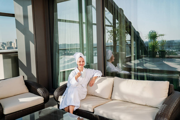 Beautiful young woman getting rest in bathrobe on terrace and drink coffee - Photo, image