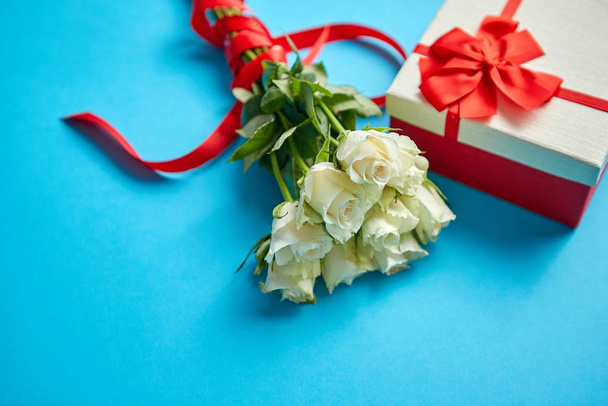 Kytice bílých růží s červenou mašlí na modrém pozadí. Zabalený dárek na straně - Fotografie, Obrázek