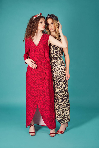Close up indoor studio fashion portrait of gorgeous women in stylish red and leopard dresses. Blonde and brunette girls isolated on turquoise background. Full-length shot of glad curly models - Photo, Image