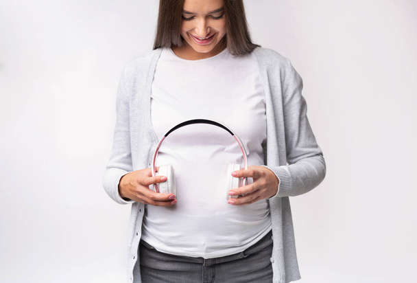 Έγκυος κορίτσι κρατώντας ακουστικά κοντά στο Belly Standing, Studio, περικοπή - Φωτογραφία, εικόνα