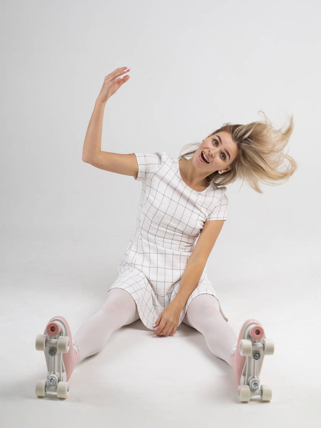 Beautiful woman shaking her head with her blonde hair flying around and sitting on a white floor in a vintage roller skates shoes - Фото, изображение