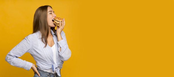 Millennial girl eating burger standing on yellow studio background, panorama - Fotó, kép