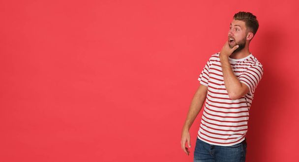 Young, characteristic man with beard with t-shirt striped looks shocked. Person isolated on red background. - Photo, Image