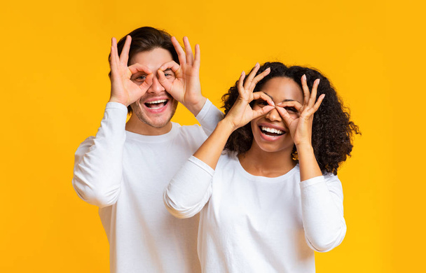 Joyful interracial couple fooling together, making funny glasses with fingers - 写真・画像