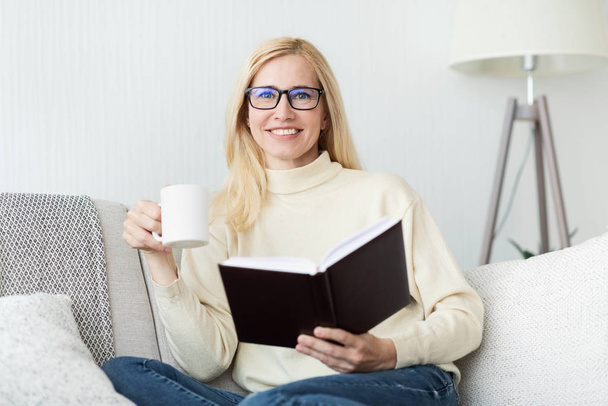 Mujer de mediana edad emocionada bebiendo té y leyendo libro
 - Foto, Imagen