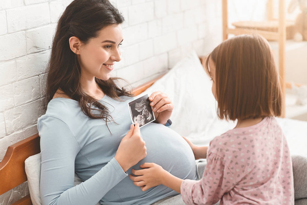 Raskaana oleva äiti näyttää ultraäänikuvauksen tyttärelleen
 - Valokuva, kuva