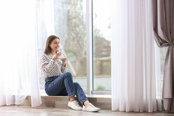 Young woman with cup near window indoors - Фото, изображение