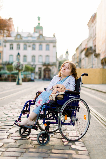 A girl in blue dress in a wheelchair outdoors in the city. The disabled person enjoys life. Young beautiful disabled woman - Foto, afbeelding