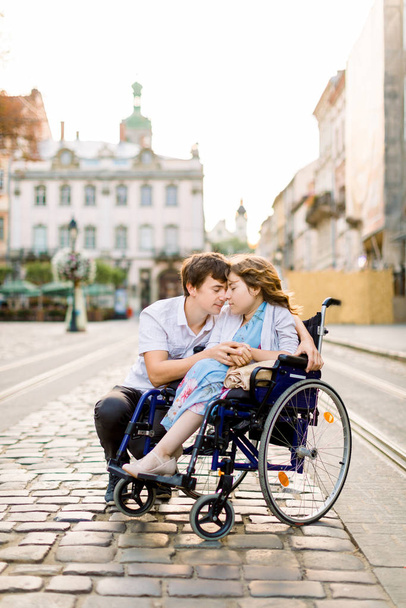 Couple in love in old city center. Pretty young girl with disease on a wheelchair and her lovely man, walking, smiling and touching foreheads, closed eyes - Foto, Bild