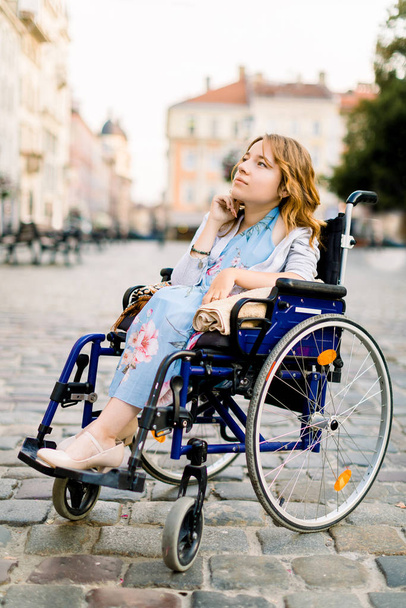 aantrekkelijke gehandicapte jonge vrouw in blauwe jurk zittend in een rolstoel, buiten in de stad - Foto, afbeelding