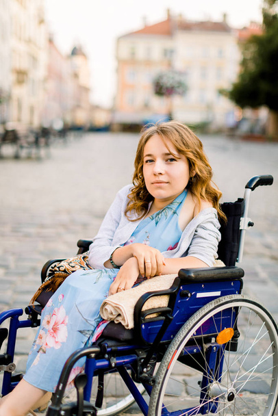 Closeup portrait of pretty young smiling disabled woman in wheelchair on the background of old city buildings - Fotó, kép