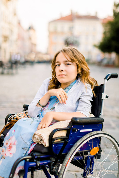 Portrait of young blond-haired woman sitting in wheelchair and looking at camera, walking outdoors in old city center - Φωτογραφία, εικόνα