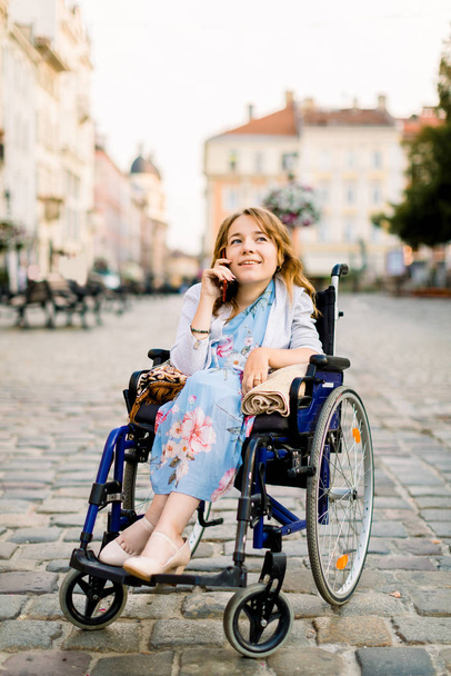 Portrait of a gorgeous young blond woman in a wheelchair enjoying her day and talking on the phone while walking in the city outdoors - Φωτογραφία, εικόνα