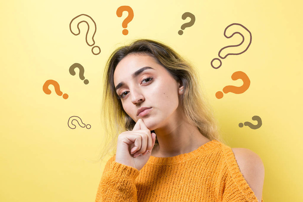 Portrait of a beautiful girl on a yellow background with question marks, looks aside with pensive expression, is considering a plan for further action. - Photo, Image