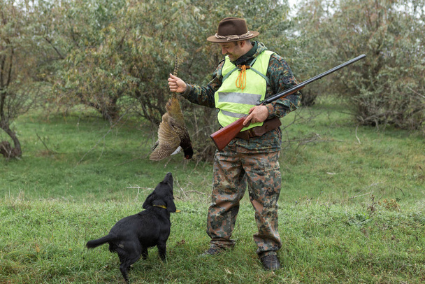 Περίοδος κυνηγιού, φθινοπωρινή περίοδος ανοιχτή. Ένας κυνηγός με ένα όπλο στα χέρια του σε ρούχα κυνηγιού στο δάσος του φθινοπώρου σε αναζήτηση ενός τροπαίου. Ένας άνθρωπος στέκεται με όπλα και κυνηγόσκυλα εντοπίζοντας το παιχνίδι. - Φωτογραφία, εικόνα