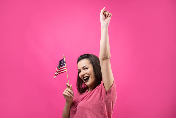 Happy young woman holding American flag against a studio pink background - Foto, Imagem
