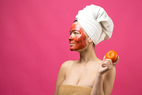 Piękny portret kobiety w białym ręczniku na głowie z czerwoną maską odżywczą na twarzy. Oczyszczanie skóry ekologiczny kosmetyczny spa zrelaksować koncepcję. Dziewczyna stoi z tyłu trzymając pomarańczowy mandarynkę. - Zdjęcie, obraz