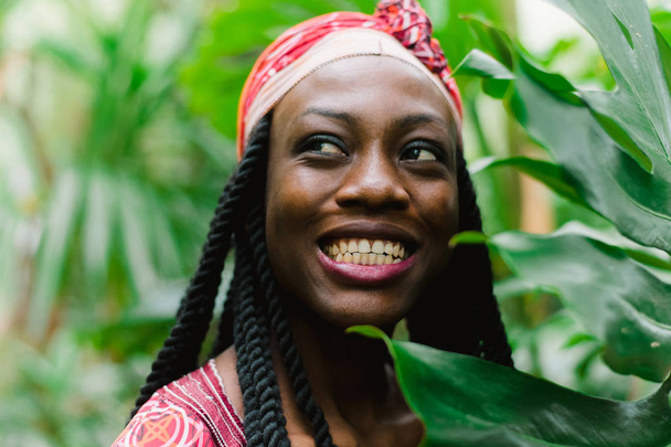 Μια όμορφη νεαρή Αφρο-Αμερικανίδα στέκεται κάτω από φοίνικες στη ζούγκλα και χαμογελά ευρέως σε ένα εθνικό αφρικανικό κοστούμι. Ευαίσθητο υπαίθριο πορτρέτο του ελκυστικού κοριτσιού από την Αφρική με σκιές ματιών - Φωτογραφία, εικόνα