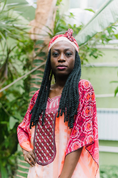 Een mooie jonge Afro-Amerikaanse vrouw staat onder palmbomen in de jungle en glimlacht breed in een nationaal Afrikaans kostuum. Gevoelig outdoor portret van het aantrekkelijke Afrikaanse meisje met oogschaduwen - Foto, afbeelding