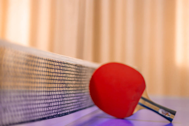 Um tênis de mesa vermelho é colocado perto da rede de uma mesa de pingue-pongue mantida dentro de casa. foco seletivo
 - Foto, Imagem