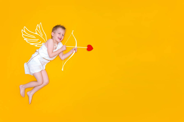 Happy smiling baby cupid in costume angel wings, bow and heart arrow isolated on yellow studio background. Copyspace for text. Valentines day concept - Fotoğraf, Görsel