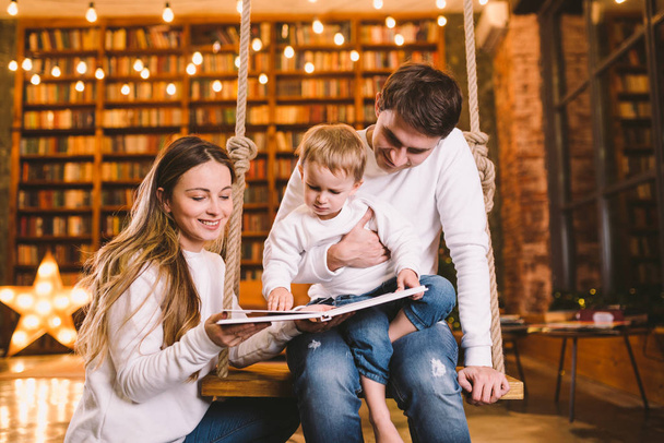 Family togetherness. Mom, dad and son reading story book together sitting on swing evening home. Family and Parenthood Concept. Happy family reads childrens book while sitting home in loft interior - Foto, Imagem