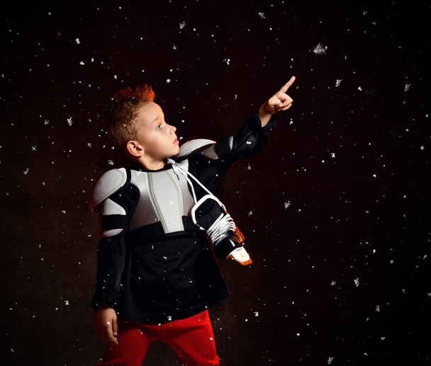 Serious boy in protective hockey uniform with skates on neck standing and reaching up over dark background with snowflakes - Fotografie, Obrázek