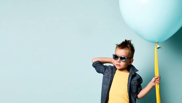 Blond boy in stylish casual clothing, sneakers and sunglasses standing near blue air balloon and looking at camera - Fotoğraf, Görsel