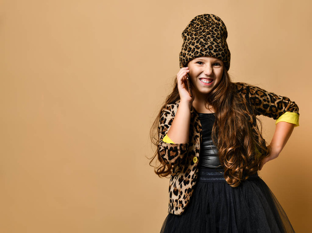 Young smiling plus size girl with long hair in leopard colored clothing and accessories standing and talking on her smartphone - Foto, afbeelding