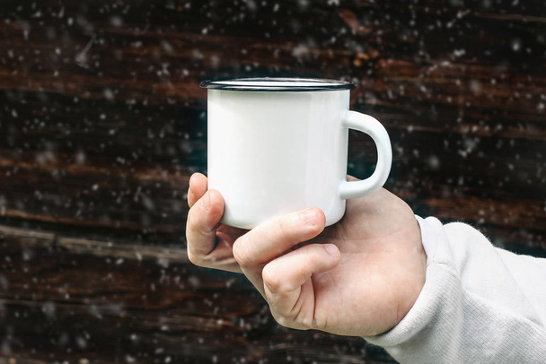 Close up of man travelers hand holding metal mug with hot drink with blurred falling snow. Outdoor tea, coffee relax time. Mockup of white enamel cup. Winter adventure and camping concept. - Fotoğraf, Görsel