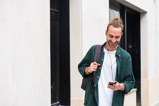 Портрет молодого человека с рюкзаком, слушающего музыку с мобильного телефона и наушников на улице города
 - Фото, изображение