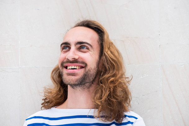 Close up portret van jonge man met baard en lang haar glimlachen en weg te kijken tegen een witte achtergrond - Foto, afbeelding