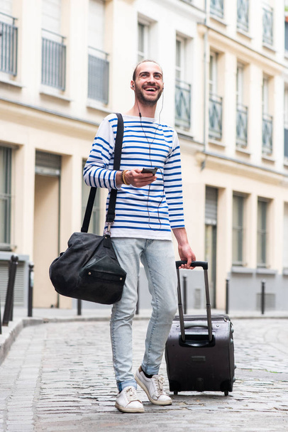 Celotělový portrét šťastného cestujícího muže kráčejícího s taškou na kufry a mobilním telefonem na ulici - Fotografie, Obrázek