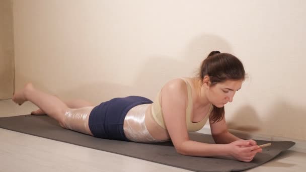 Uma mulher está deitada num tapete de ginástica com um telefone. Coxas e barriga embrulhadas em plástico
 - Filmagem, Vídeo