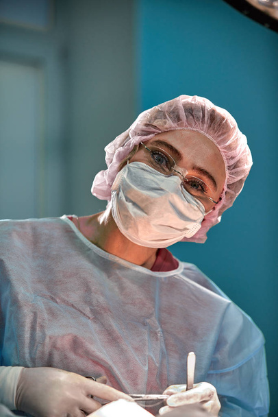 Close up portrait of young female surgeon doctor wearing protective mask and hat during the operation. Healthcare, medical education, surgery concept - Photo, Image