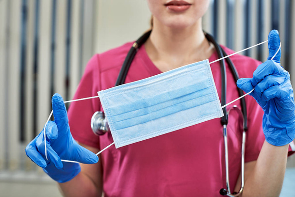 jovem mulher bonita médico em uniforme rosa e máscara em suas mãos fechar no fundo da clínica
 - Foto, Imagem