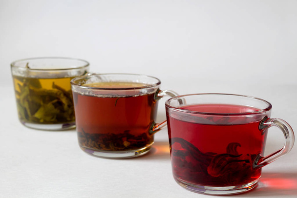 Hot drinks. Healthy lifestyle. Three glass cups with black, red and green tea on a white background. Red hot Hibiscus tea. Green hot tea. Black hot tea. - Photo, Image