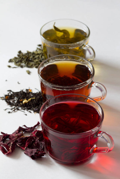 Three glass cups with black, red and green tea on a white background. Red hot Hibiscus tea with dry flowers. Healthy lifestyle.  Green hot tea. Black hot tea. Hot drinks.Vertical orientation.top view - Photo, Image