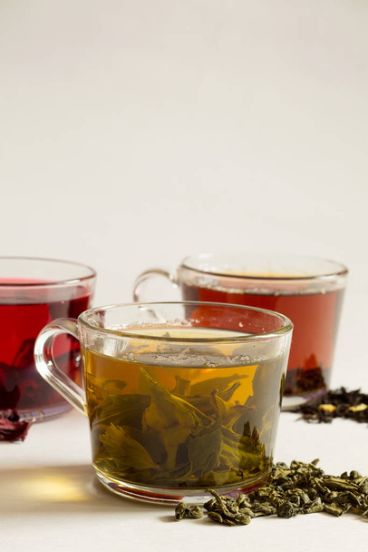 Green hot tea with dry leaves on a white. Three glass cups with black, red and green tea on a white background. Red hot Hibiscus tea. Black hot tea. Healthy lifestyle.Hot drinks. vertical orientation - Photo, image