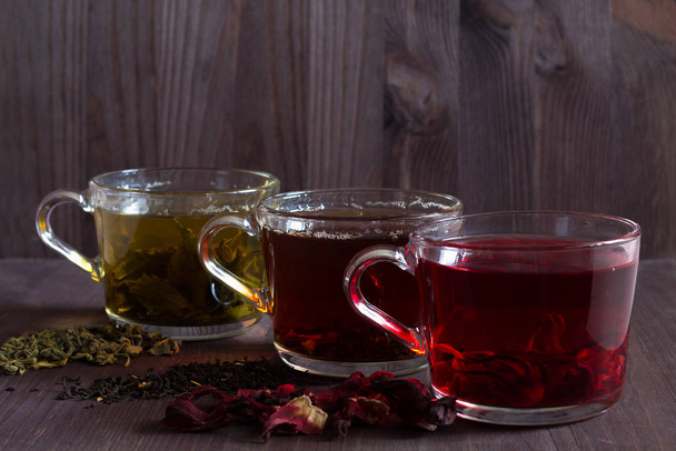 Три стакана черного, красного и зеленого чая с сухими листьями и цветами на темном деревянном фоне. Горячий чай с гибискусом. Черный горячий чай. Зеленый горячий чай на деревянном столе
. - Фото, изображение