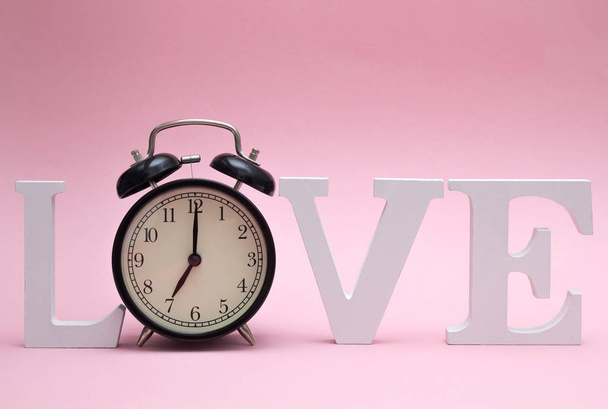 Λέξη Αγάπη φτιαγμένη από ξύλινα λευκά γράμματα και μαύρο ξυπνητήρι αντί για το γράμμα Ο σε ροζ φόντο. Ημέρα του Αγίου Βαλεντίνου. Ώρα για αγάπη - Φωτογραφία, εικόνα