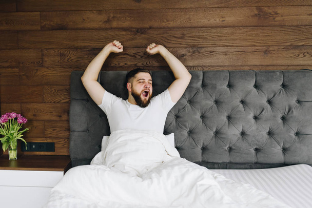 Молодой кавказский бородатый мужчина зевает и растягивается в своей яркой современной спальне утром
  - Фото, изображение